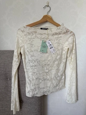 Vit spetsblus från Gina Tricot - Säljer en elegant vit spetsblus från Gina Tricot med lång ärm och blommigt mönster. Tillverkad av återvunnen polyester och har en figurnära passform. Perfekt för dig som vill ha en romantisk och trendig look.