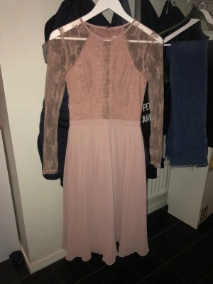 Ljusrosa långklänning med spetsärmar - Elegant ljusrosa långklänning med långärmade spetsdetaljer och rund halsringning.  plisserad i ett mjukt, flytande tyg. Dragkedja bak för enkel påklädning. Perfekt för dig som vill ha en romantisk look.