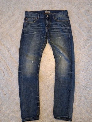 Blå slim fit jeans med slitningar - Snygga blå slim fit jeans med slitningar och tvättade detaljer. Jeansen har en modern passform och är perfekta för en trendig vardagslook. !Trasig dragkedja! Snygg gul lapp där bak. W30 l32