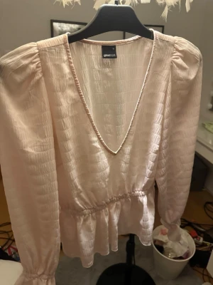 Ljusrosa blus från Gina Tricot - Söt ljusrosa blus från Gina Tricot med v-ringning och puffärmar. Blusen har ett lätt glansigt, strukturerat tyg och smock i midjan som ger en fin siluett. Perfekt att styla med jeans eller kjol för en trendig look.