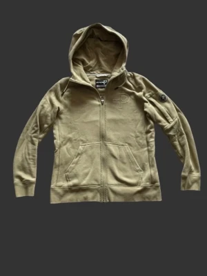 Pelle P Zip Hoodie - Snygg beige hoodie från Pelle P i storlek Mmed dragkedja och huva. Har två stora fickor framtill och diskret broderad logga på bröstet samt patch på ärmen. Tillverkad i mjukt bomullsmaterial, perfekt för chill dagar eller när du vill ha en clean look. Fråga gärna om du undrar något. Priset är inte fast!