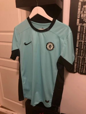 Chelsea ljusblå fotbollströja Nike - Chelsea FC fotbollströja i ljusblått med svarta detaljer och svart Nike-logga. Tröjan har klubbmärke på bröstet, rund hals och korta ärmar. Perfekt för träning eller match. Tillverkad i lätt och ventilerande material.