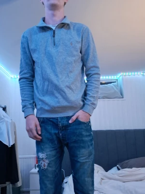 Grå halvzip från Polo Ralph Lauren - Snygg grå halvzip-tröja från Polo Ralph Lauren. Tröjan har hög krage med dragkedja och ribbade muddar vid ärmslut och nederkant. Perfekt till jeans för en clean och stilren look.