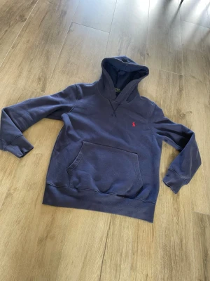 Mörkblå polo Ralph lauren hoddie med röd logga - Säljer en mörkblå hoodie med huva och känguruficka framtill. Tröjan har en liten röd broderad logga på bröstet och ribbade muddar vid ärmslut och nederkant. Perfekt för en avslappnad och stilren look.