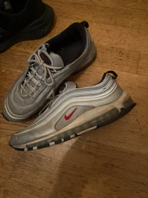 Nike Air Max 97 silver sneakers - Säljer ett par Nike Air Max 97 sneakers i silver med reflekterande detaljer och röd swoosh på sidan. Skorna har en vågig design, synlig luftdämpning i sulan och vita samt svarta inslag. Klassisk snörning och låg profil.