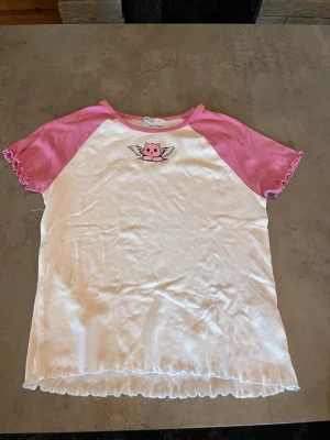 Söt t-shirt med rosa ärmar och björn - T-shirt med vita framsida och baksida samt rosa raglanärmar. På bröstet finns ett tryck av en rosa björn med vingar. Ärmsluten och nederkanten har volangdetaljer. Perfekt för en lekfull och gullig stil.