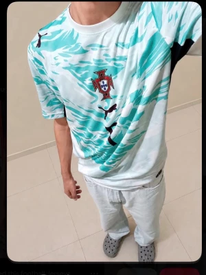 Portugal t-shirt med turkost mönster - Säljer en Portugal t-shirt med turkost och vitt abstrakt mönster samt svart Puma-logga. T-shirten har rund hals och korta ärmar. Perfekt för dig som vill sticka ut med en sportig look.