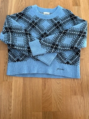 Blå stickad tröja från Ganni - Supersnygg blå stickad tröja från Ganni med svart och vitt rutmönster. Tröjan har ribbstickade muddar och nederkant samt broderad Ganni-logga framtill. Perfekt oversized fit och mjukt material som känns lyxigt.