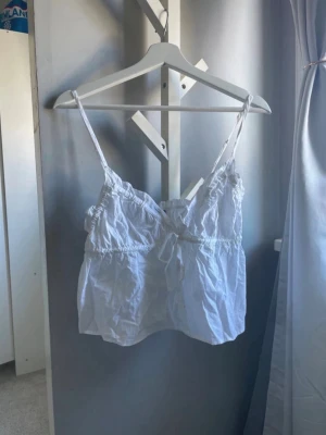Vit linnetopp med knyt från Gina Tricot - Söt vit linnetopp från Gina Tricot med tunna axelband, v-ringning och volangdetaljer. Toppen har en knytning framtill och är lätt utsvängd nedtill. Perfekt för varma dagar och enkel att styla med jeans eller kjol.