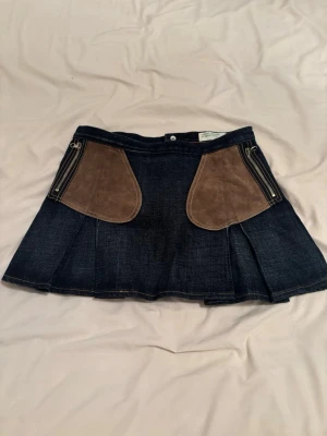 Kort jeanskjol med mockadetaljer - Snygg kort jeanskjol med bruna mockadetaljer och dragkedjor framtill. Kjolen har en A-linjeformad siluett och markerade sömmar som ger en cool look. Perfekt för dig som vill ha en unik twist på din outfit.
