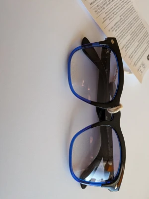 Ray-Ban blå och svarta solglasögon - Säljer ett par Ray-Ban solglasögon med svart ovandel och blå transparent nederdel på bågarna. Glasen är lätt tonade och bågarna har en klassisk, fyrkantig form. Snygga detaljer med metallnitar vid hörnen. Perfekta för dig som vill sticka ut lite extra.