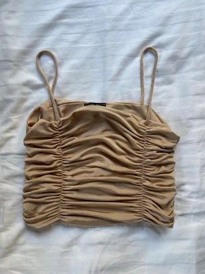 Beige ruched linnetopp med smala band - Trendig beige linnetopp med smala axelband och snygga rynkade detaljer framtill och baktill. Toppen är croppad och har en tight passform som framhäver siluetten. Perfekt för att styla med jeans eller kjol för en cool look.