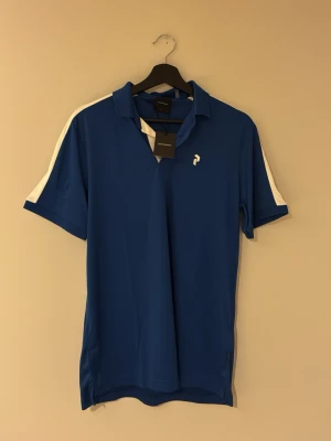 Helt oanvänd peak performance golfpike, tröjan har lappar kvar.  - Snygg blå golfpikétopp med vit rand längs axlarna och ett litet vitt tryck på bröstet. Klassisk krage och korta ärmar. Perfekt för dig som gillar sportig stil. Oanvänd tröja!! Lappar kvar. 