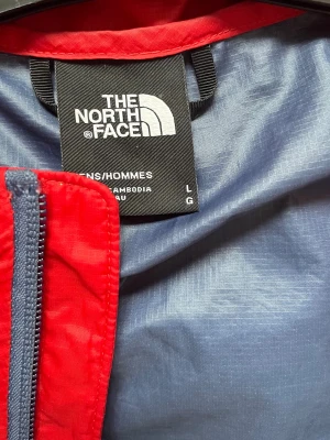 Röd vindjacka från The North Face - Snygg röd vindjacka från The North Face med marinblå detaljer upptill och på huvan. Jackan har hel dragkedja framtill, två sidofickor med dragkedja och justerbar huva. Tillverkad i lätt och slitstarkt syntetmaterial, perfekt för blåsiga dagar.