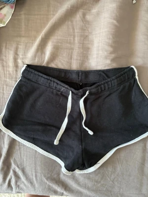 Svarta sportiga shorts med snörning - Svarta sportiga shorts med vit snörning i midjan och vita kanter. Klassisk retrostil med rundad nederkant och elastisk midja. Perfekta för sommarens alla aktiviteter.