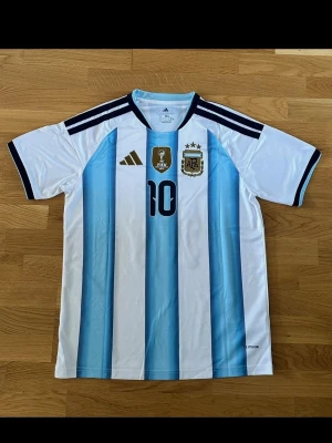 Argentina Hemmatröja - Helt ny och oanvänd Argentina-hemmatröja med Messi 10 på ryggen.  Skick: Helt ny med lappar kvar (NWT)  Storlek: Large (L) – Slim Fit  Märken: Tre stjärnor + officiellt FIFA World Champions 2022-märke på bröstet  Märke: adidas (AEROREADY) 