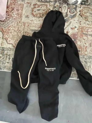 Svart Essentials Fear of God set - Säljer ett svart loungewear set från Essentials Fear of God. Setet består av en hoodie med huva och ett par matchande byxor med vit dragsko. Båda plaggen har loggan tryckt i vitt på framsidan. Perfekt för chill dagar hemma.