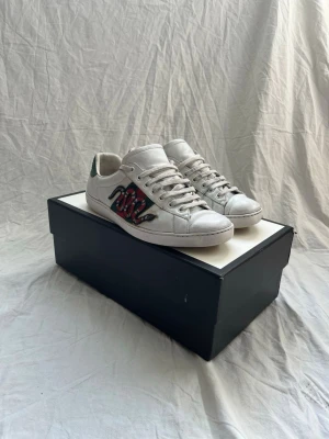 Vita låga sneakers med orm-detalj - Säljer ett par vita låga sneakers med cool orm-detalj i rött och svart på sidan. Skorna har klassisk snörning och är tillverkade i läder med en grön detalj på hälen. Perfekta för dig som vill sticka ut med din stil.