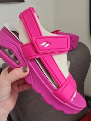 Helt nya rosa sandaler från Skechers - Säljer ett par knallrosa sandaler, stl 37, från Skechers med chunky sula och justerbara kardborreband. Sandalerna har en sportig design med synlig luftbubbla i sulan och detaljer i vitt. Perfekta för dig som vill sticka ut med färg och komfort. Helt nya och oanvända!
