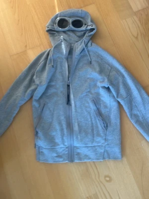 Cp company hoodie - Säljer en grå hoodie med dragkedja och två fickor framtill. Huvan har inbyggda glasögon och dragsko för justering. Hoodien är tillverkad i mjukt material och har en modern, sportig look. Perfekt för dig som vill sticka ut med en unik detalj på huvan.Hoodien är i XS men passar även storlek S