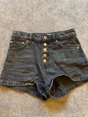 Svarta högmidjade jeansshorts - Snygga svarta högmidjade jeansshorts med fem knappar framtill och klassiska fickor både fram och bak. Shortsen har bälteshällor och är gjorda i ett slitstarkt bomullsmaterial. Perfekta för varma dagar och en avslappnad stil.