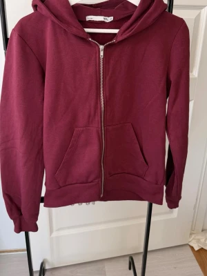 Vinröd hoodie med dragkedja - Säljer en vinröd hoodie med dragkedja framtill och två fickor. Hoodien har ribbade muddar vid ärmslut och nederkant samt en klassisk huva. Perfekt för chill dagar och enkel att matcha med jeans eller mjukisbyxor.
