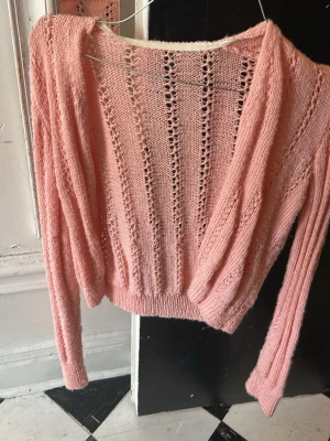 Rosa stickad omlottkofta - Söt rosa stickad omlottkofta med långa ärmar och hålmönster längs hela plagget. Kortare modell med ribbstickad nederkant och ärmslut. Perfekt att slänga över en topp för en trendig lager-på-lager-look.