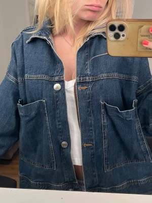 Oversized blå jeansjacka - Säljer en oversized blå jeansjacka i klassisk denim med stora framfickor och silverfärgade knappar. Jackan har en avslappnad passform, krage och långa ärmar. 