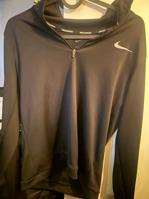 Svart löpartröja med halv dragkedja Nike - Svart långärmad löpartröja från Nike med halv dragkedja framtill och hög krage. Tillverkad i ett lätt, stretchigt funktionsmaterial. Reflekterande Nike-logga på bröstet och reflexdetalj baktill. Perfekt för löpning och träning.
