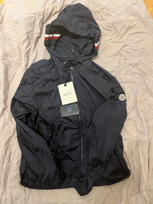 Marinblå vindjacka från Moncler - Snygg marinblå vindjacka från Moncler med huva och dragkedja framtill. Jackan har Moncler-logga på ärmen och vita samt röda ränder på huvan. Tillverkad i lätt, glansigt material och har justerbar dragsko vid huvan. Perfekt för en sportig och trendig look.