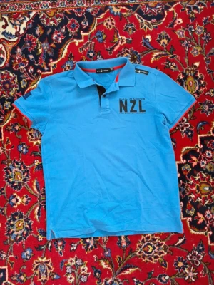 Blå Sail Racing NZL t-shirt - Blå t-shirt från Sail Racing med NZL-tryck på bröstet och Nya Zeelands flagga på ärmen. Klassisk pikékrage med knappar och röda detaljer på insidan av kragen och ärmsluten. Tillverkad i bomull och har normal passform.