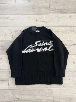 Svart stickad Saint Laurent-tröja - Svart stickad tröja från Saint Laurent med vit, fluffig logotyp i graffiti-stil över bröstet. Tröjan har rund halsringning, långa ärmar och breda ribbade muddar vid ärmslut och nederkant. Perfekt för dig som gillar streetwear och exklusiva märken.