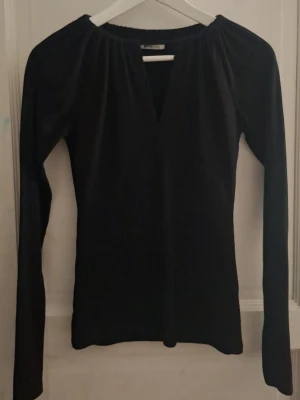 Svart långärmad topp med v-ringning - En stilren svart långärmad topp med djup v-ringning och lätt rynkad halsringning. Toppen har en smickrande passform och är perfekt att matcha med både jeans och kjol. Materialet känns mjukt och följsamt.