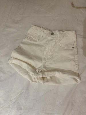 Vita högmidjade jeansshorts - Säljer ett par vita högmidjade jeansshorts med uppvikta ben och klassisk femficksdesign. Perfekta för varma dagar och enkla att matcha med olika toppar. Stängs med knapp och dragkedja framtill.