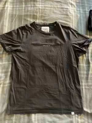 Mörkgrå t-shirt från Les Deux - Säljer en stilren mörkgrå t-shirt från Les Deux med diskret logga på bröstet. Klassisk passform och rund halsringning. Tillverkad i mjuk bomull som känns skön mot huden. Perfekt till jeans eller shorts.