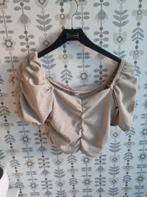 Beige manchester topp med puffärm - Trendig beige topp i manchester med puffiga korta ärmar och söt omlott-look framtill. Toppen har en croppad passform och elastiska axelband som ger en offshoulder-stil. Perfekt för dig som vill sticka ut med en unik topp.