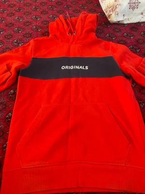 Röd hoodie från Jack & Jones - Snygg röd hoodie från Jack & Jones Originals med svart panel och vit text på bröstet. Klassisk känguruficka framtill och justerbar huva med snören. Perfekt för en avslappnad streetwear-look.