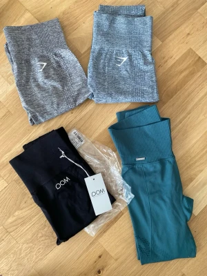Träningsleggings i olika färger- strl xs - Säljer flera par träningsleggings i olika färger och material. Två par är ljusgrå med mönster och logga, ett par är svart med hög midja och ett par är petrolblå med ribbade detaljer och meshpartier. Perfekta för gymmet eller löpning. Alla är strl Xs, endast testade 