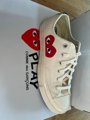 Converse x Comme des Garçons Play låga - Säljer ett par Converse x Comme des Garçons Play sneakers i offwhite canvas med den ikoniska röda hjärtloggan på sidan. Skorna har vita snören, metallöljetter och klassisk gummisula. Perfekta för dig som vill ha en clean men ändå unik look.