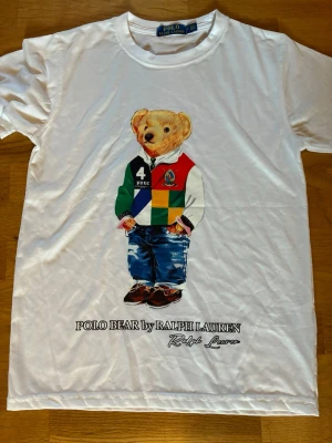 Vit Polo Bear t-shirt från Ralph Lauren - Vit t-shirt med ikoniskt Polo Bear-tryck från Ralph Lauren. Björnen har färgglada kläder och jeans, och under motivet står det 'POLO BEAR by RALPH LAUREN'. Klassisk rund hals och korta ärmar. Perfekt statement-plagg för dig som gillar streetstyle.             Kontakta gärna vid frågor och prisförslag. Skickar inom 24h