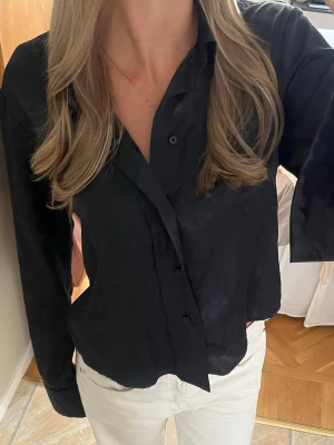 HOPE skjorta - Säljer en svart skjorta med lång ärm och klassisk krage. Skjortan har knappar framtill och en snygg satinliknande finish som ger en stilren vibe. Perfekt att styla till både jeans och kjol för en trendig look.