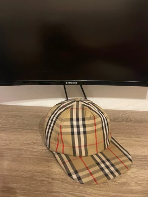 Burberry beige keps - Säljer en klassisk Burberry keps i beige med det ikoniska rutmönstret i svart, vitt och rött. Justerbar baktill med svart plastspänne och Burberry London England-märke. Säljer för att den ej används längre skruv vid frågor pris kan diskuteras!