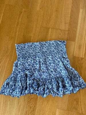 Blå blommig volangkjol - Söt blå kortkjol med vitt blommönster och volangkant. Kjolen har smock i midjan för en bekväm passform och är perfekt för varma dagar. Tillverkad i ett lätt och luftigt material som känns skönt mot huden.