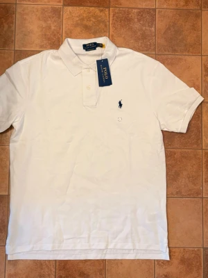 Polo Ralph Lauren piké – L - Helt ny Polo Ralph Lauren pikétröja i klassiskt vitt. Aldrig använd och kommer med originaltags kvar. Stilren modell med diskret marinblå Polo Ralph Lauren-logga på bröstet. 