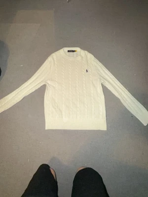 Vit kabelstickad tröja från Polo Ralph Lauren - Kabelstickad vit tröja från Polo Ralph Lauren med rund halsringning och den klassiska broderade loggan på bröstet. Tröjan har långa ärmar och ribbade muddar vid ärmslut och nederkant. Perfekt för en clean och stilren look. Obs kvitto finns ej då den e köpt utomlands