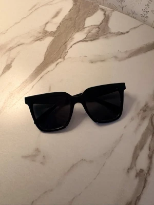 Svarta kantiga solglasögon - Snygga svarta solglasögon med breda bågar och kantig form. Glasen är mörka och ramen är i plast, vilket ger en modern och stilren look. Perfekta för att lyfta vilken outfit som helst.