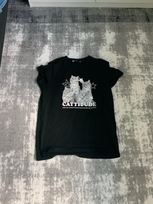 Svart t-shirt med kattmotiv - Cool svart t-shirt med tryck av två katter och texten 'Cattitude' framtill. T-shirten har korta ärmar och en klassisk rund hals. Perfekt för dig som gillar katter och vill ha en avslappnad stil.