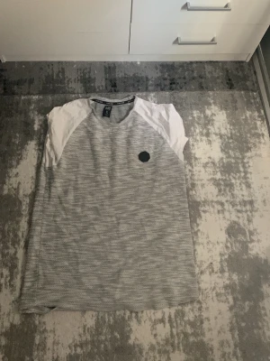 Gråvit randig t-shirt med raglanärm - Säljer en gråvit t-shirt med tunna horisontella ränder och vita raglanärmar. T-shirten har en rund halsringning och en liten svart patch på bröstet. Perfekt för en avslappnad stil.