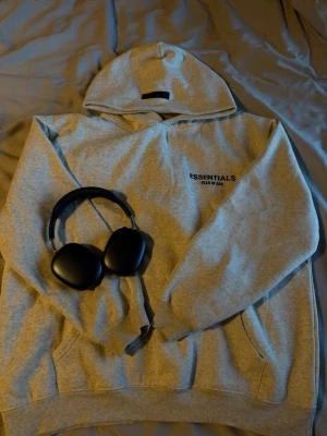  Essentials hoodie - Snygg grå hoodie från Essentials Fear of God med svart logga på bröstet. Klassisk passform med huva och känguruficka framtill. Tillverkad i mjukt bomullsmaterial som är perfekt för en avslappnad streetwear-look. Passar även L