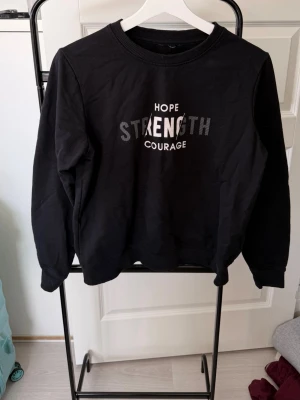Svart sweatshirt med tryck - Cool svart sweatshirt med texten 'HOPE STRENGTH COURAGE' i vitt och grått framtill. Tröjan har rund halsringning, långa ärmar och ribbade muddar vid ärmslut och nederkant. Perfekt till jeans eller mjukisbyxor.
